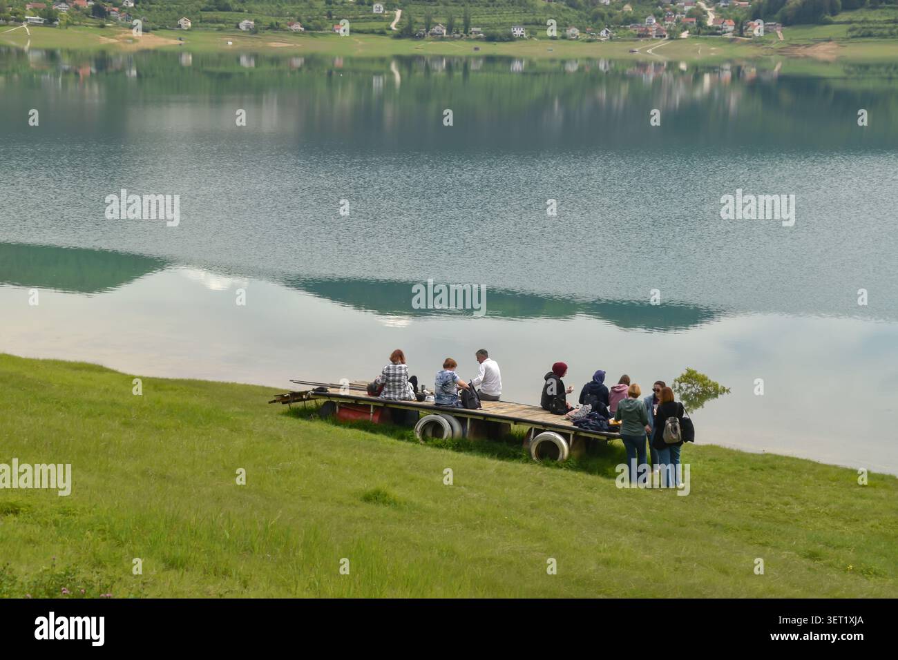 Auf einem Holzrailer am ruhigen, reflektierenden Wasser des Rama Lake, umgeben von grasbewachsenen Hügeln, treffen sich verschiedene Freunde. Stockfoto