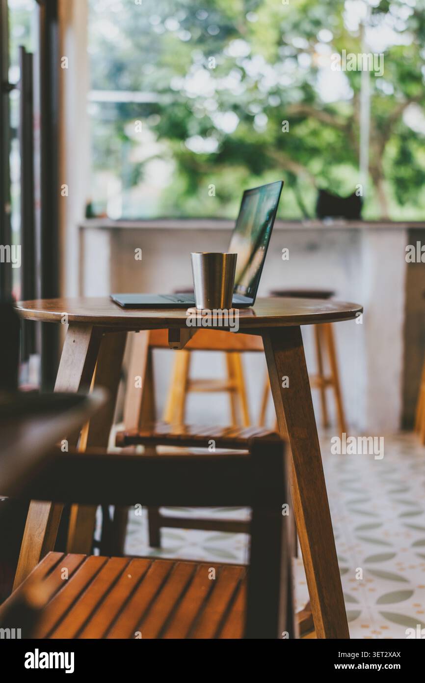 Gemütliche Café-Ecke in Bangkok, Thailand - Holztisch mit Laptop und Metallbecher, weiches Tageslicht Stockfoto