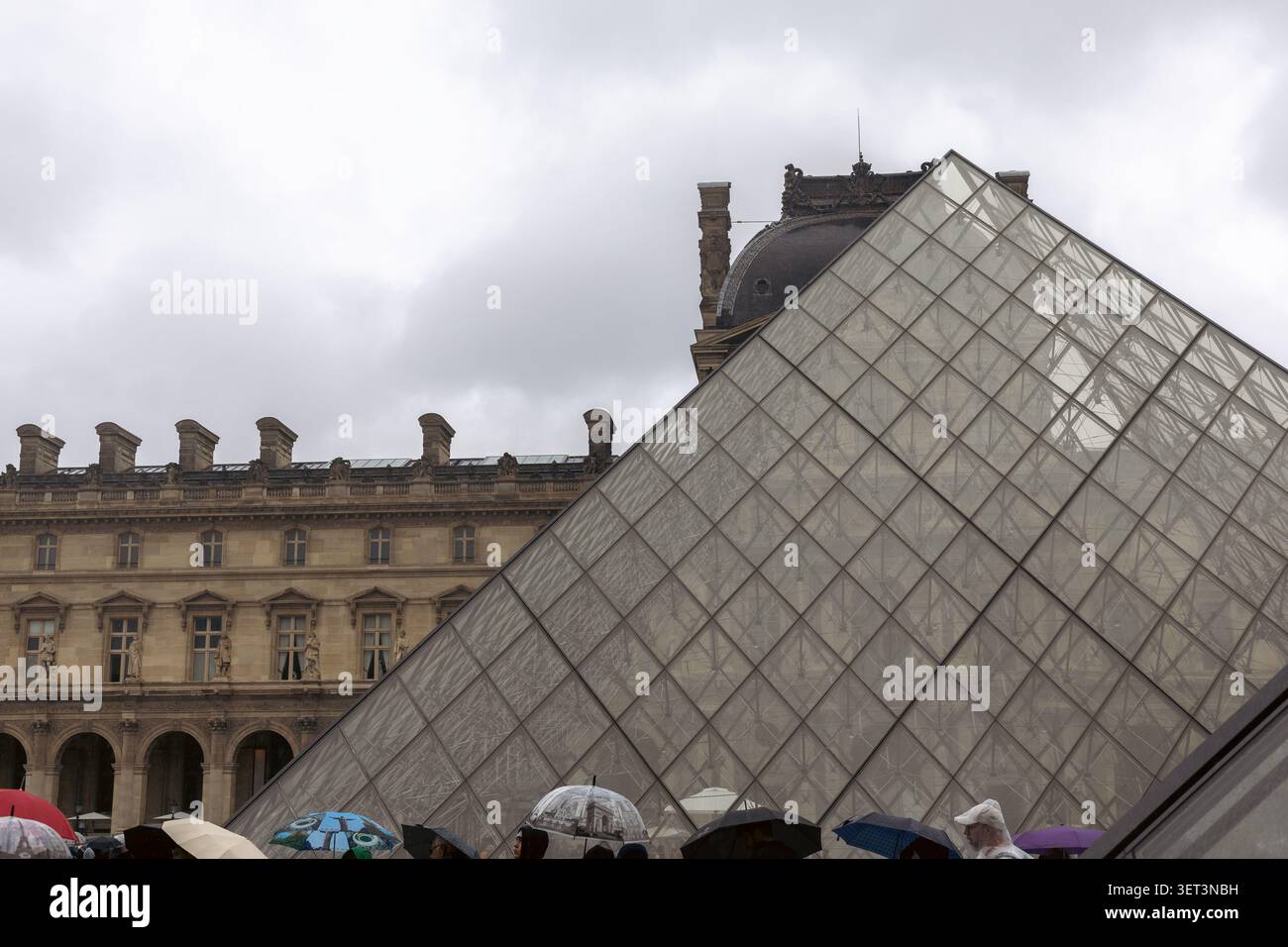 Paris, Frankreich - 3. Mai 2026: Die Menschen stehen mit Regenschirmen beim Besuch des Louvre-Museums in Paris. Die Glaspyramide ist vor einem bewölkten SK sichtbar Stockfoto