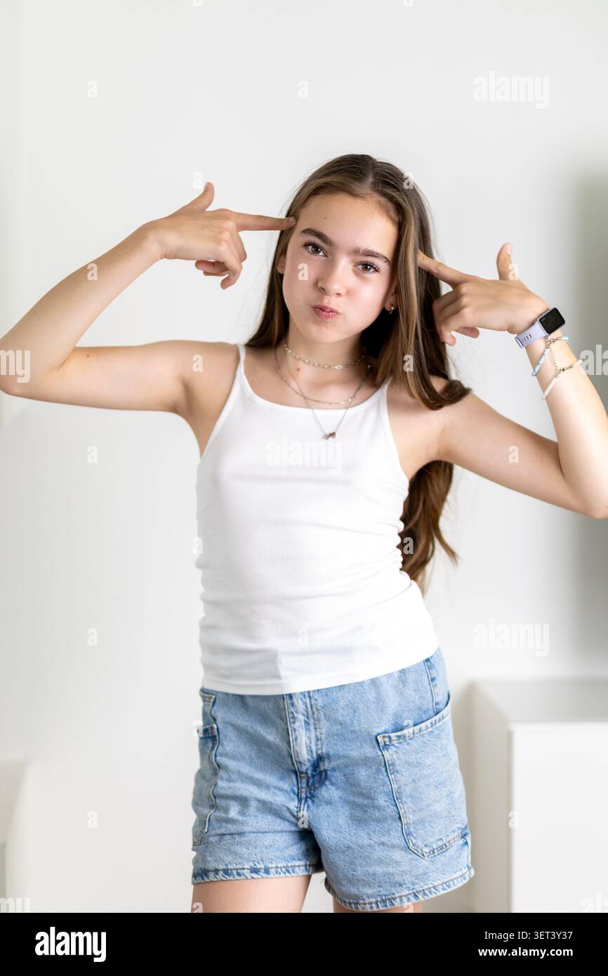 Ein Mädchen steht vor einer weißen Wand und zeigt ihre verspielte Seite. Sie trägt ein weißes Tanktop und Jeansshorts. Ihre Finger zeigen auf ihren Kopf und fügen hinzu Stockfoto