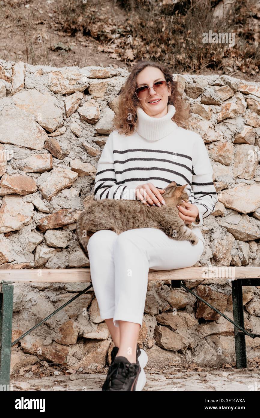 Frau Katze streichelt eine stilvolle Frau sitzt auf einer Bank und streichelt eine Tabbykatze vor einer rustikalen Steinmauer draußen Stockfoto