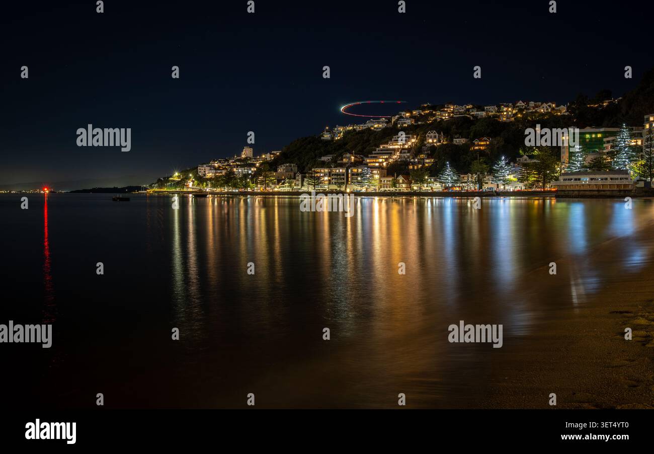 Langzeitfoto von Oriental Bay, Wellington, mit glitzernden Stadtlichtern und Hafenreflektionen. Die leichte Spur des Flugzeugs sorgt für dynamische Bewegungsabläufe. Stockfoto