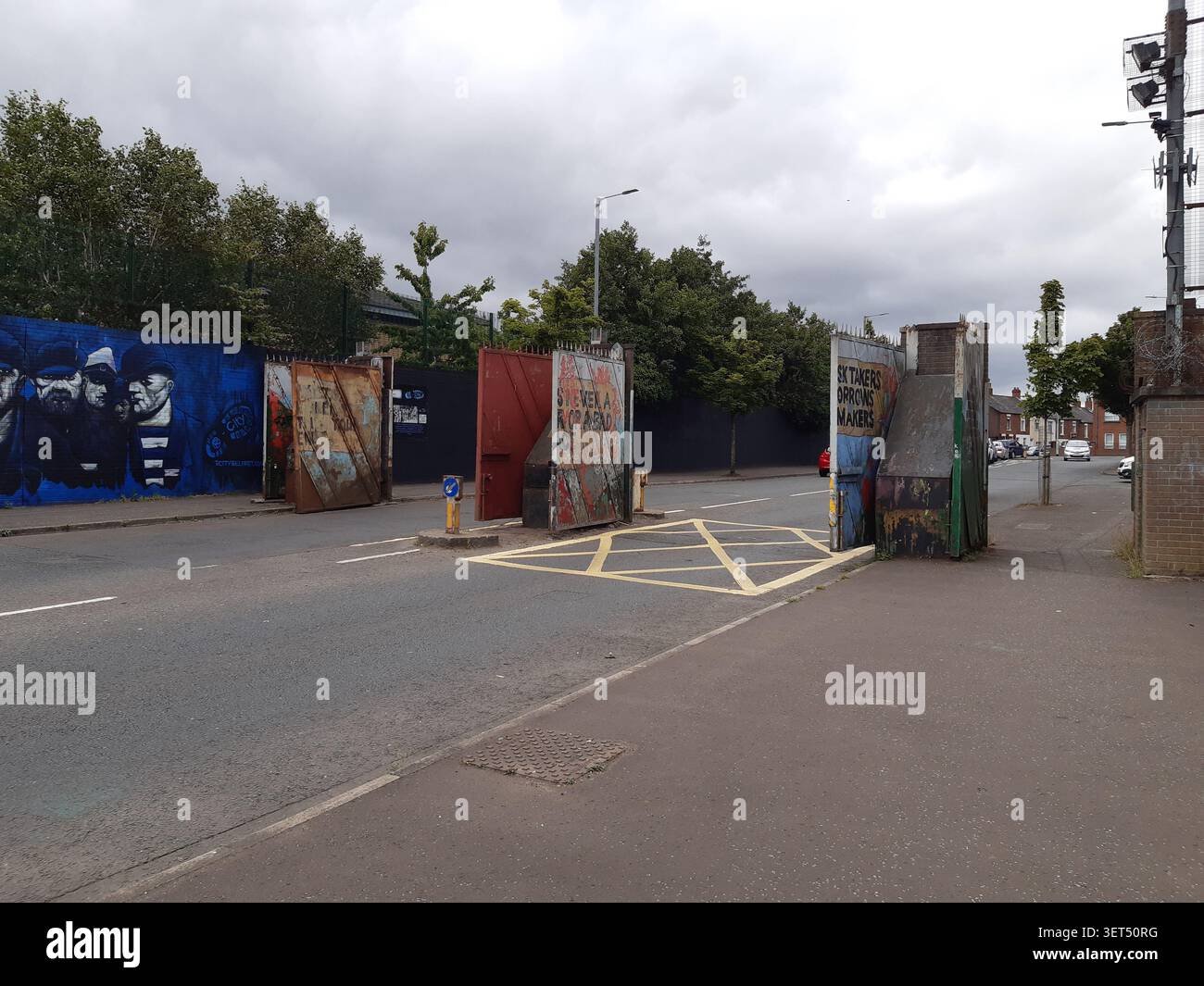 Straßenblick auf die Friedensmauer und die Sicherheitstore in Belfast, mit lokalen Wandgemälden und städtischer Infrastruktur an einer ruhigen Stadtdurchfahrt. Stockfoto