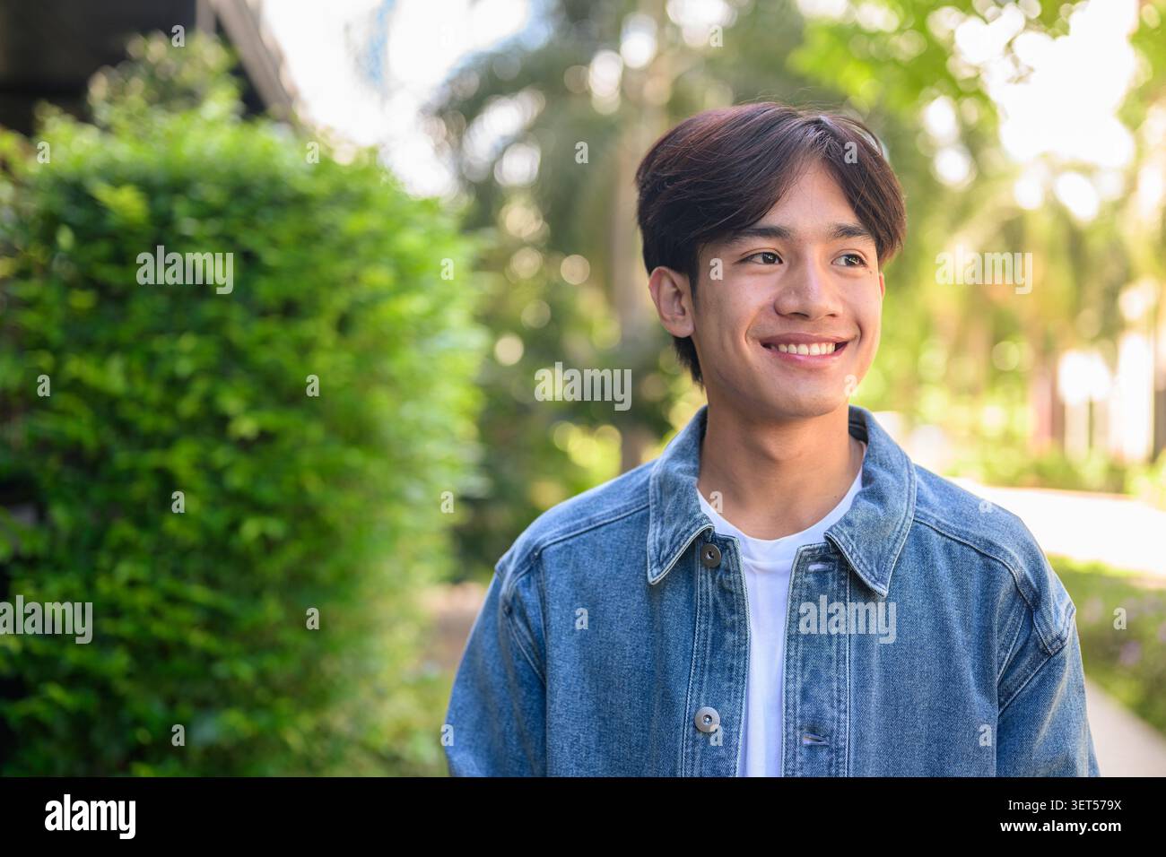 Horizontales mittleres Porträt eines fröhlich lächelnden jungen südostasiatischen Thai-Isan-Mannes in einer Jeansjacke, der mit grünem Bokeh nach draußen blickt Stockfoto