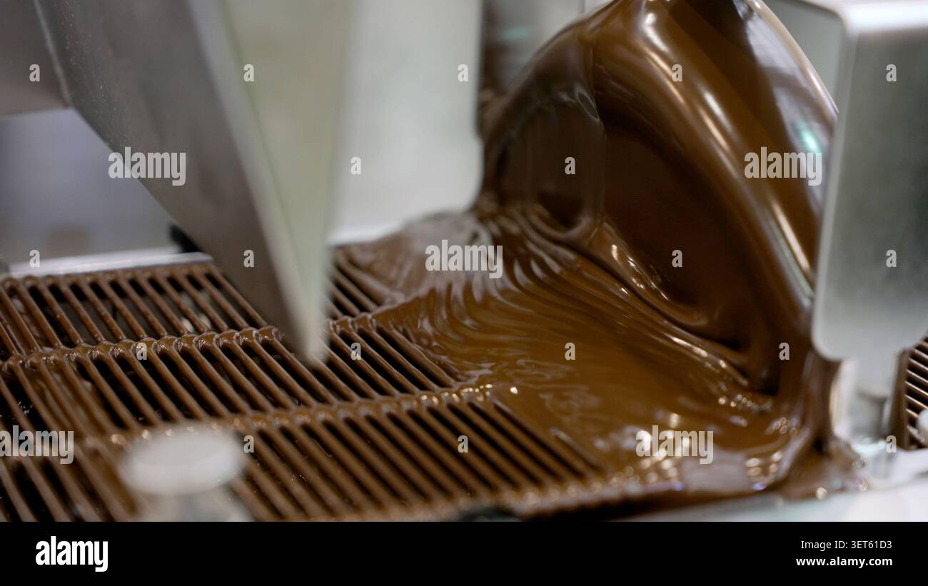 Geschmolzene Schokolade, die in einer Schokoladenfabrik von einer Schokoladentemperaturmaschine auf ein Kühlregal gegossen wird Stockfoto