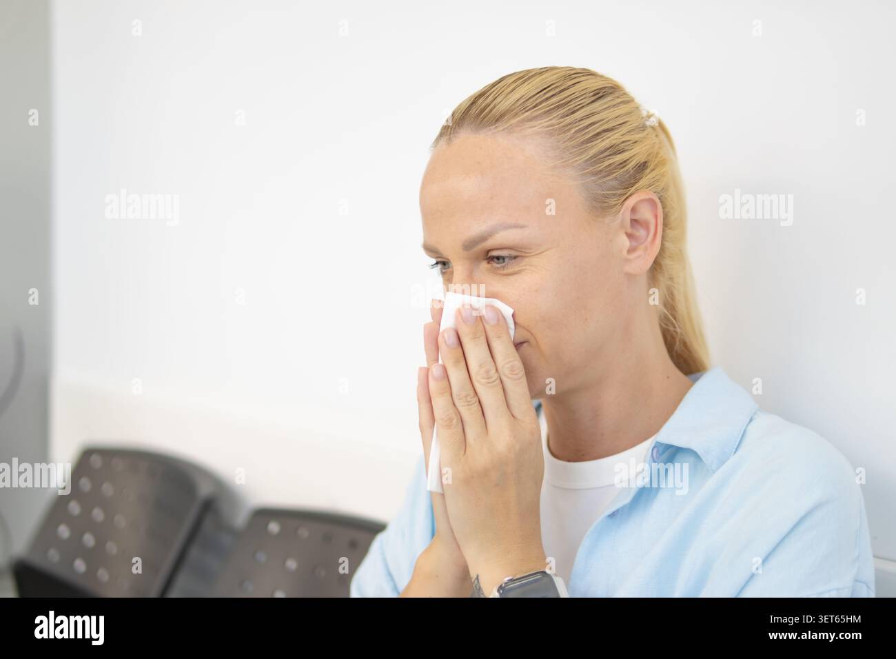 Frau, die in einem Wartezimmer sitzt, ihre Nase in ein Papiertuch bläst, was darauf hinweist, dass sie sich krank fühlt wegen einer Erkältung, Grippe oder saisonalen Allergien, exper Stockfoto