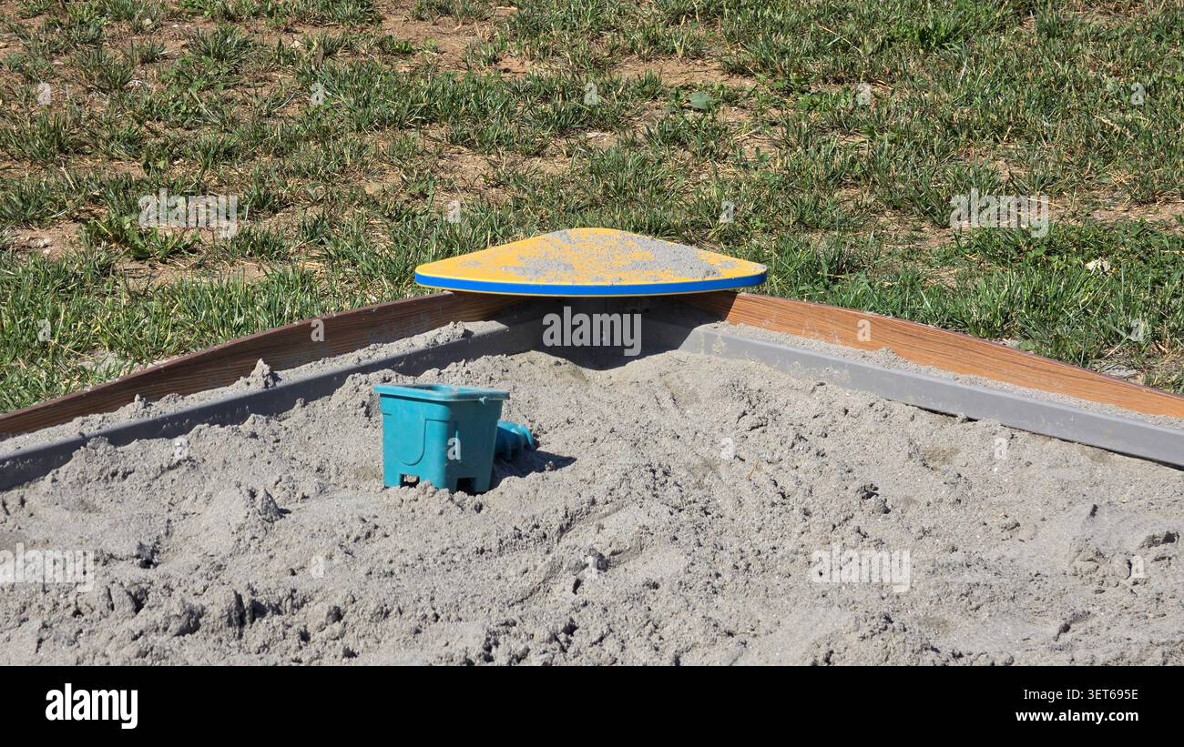 Eine dreieckige gelbe Eckbank sitzt am Holzrand eines öffentlichen Spielplatzes, gefüllt mit grauem Spielsand und einem kleinen blauen Plastikeimer Stockfoto