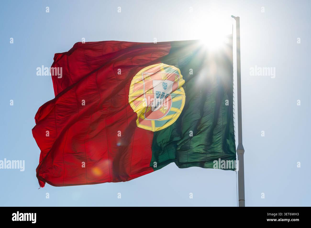 Große portugiesische Flagge, die am Fahnenmast winkt, mit strahlendem Sonnenlicht am blauen Himmel Stockfoto