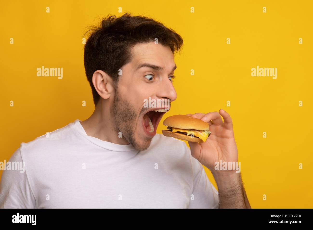 Aufgeregter Junger Mann, Der Burger Mit Freudvollem Ausdruck Hält, Gelber Hintergrund, Fast Food-Konzept Stockfoto