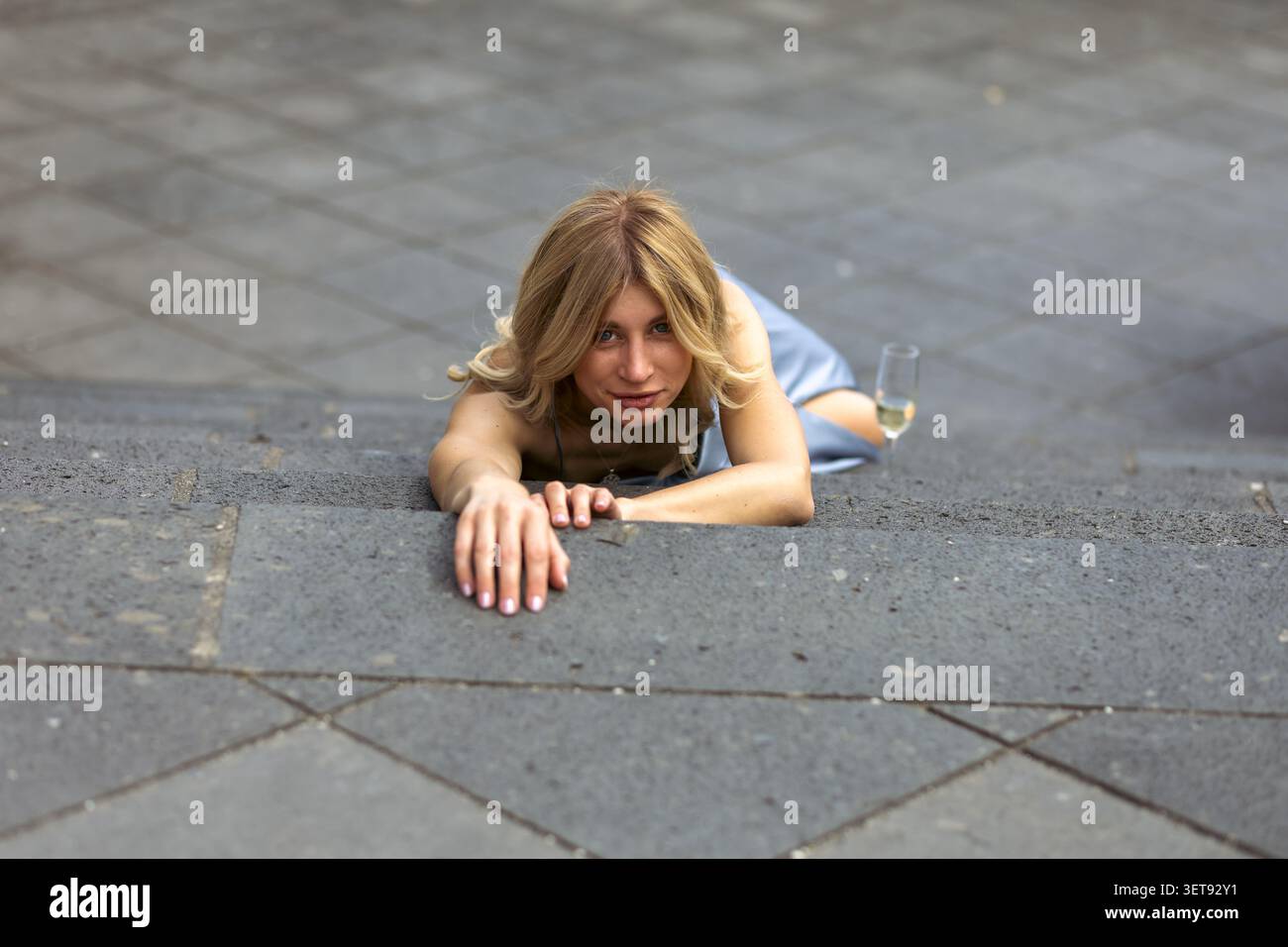 Eine Frau ruht auf einer Treppe, während sie ein Glas hält. Sie scheint einen Moment bei einem gesellschaftlichen Ereignis zu genießen. Die Sonne scheint, während die Leute in der Nähe reden Stockfoto