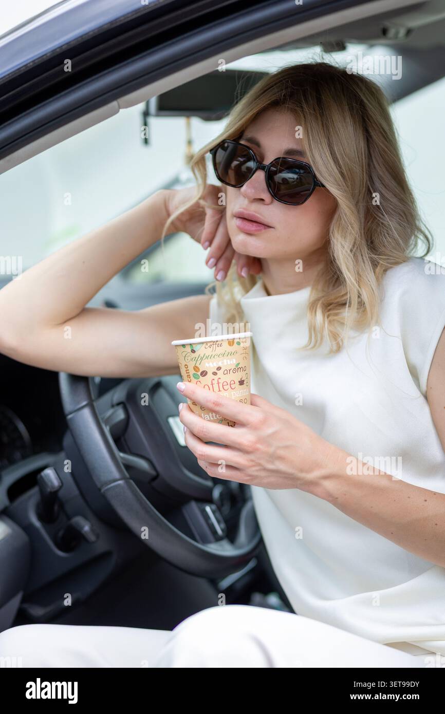 Eine Frau sitzt mit offener Tür in einem Auto. Sie hält eine Kaffeetasse in einer Hand und scheint zu denken. Die Sonne scheint, und sie sieht entspannt aus Stockfoto