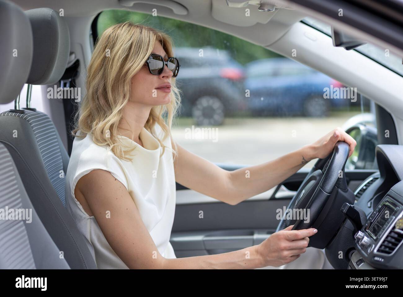 Eine Frau sitzt in einem Auto, hält das Lenkrad und blickt aus dem Fenster. Sie trägt eine Sonnenbrille und konzentriert sich auf die Straße. Die Szene nehmen Stockfoto