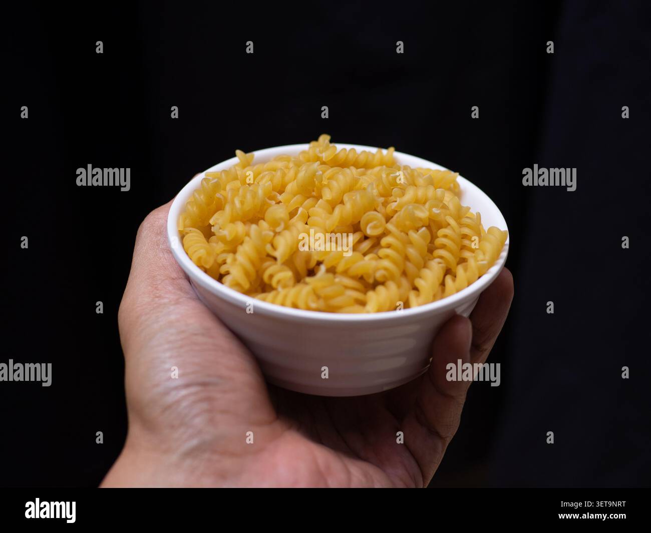 Rohe, trockene Fusilli-Nudeln auf einer weißen Schüssel, die von der linken Hand auf schwarzem Hintergrund gehalten wird Stockfoto