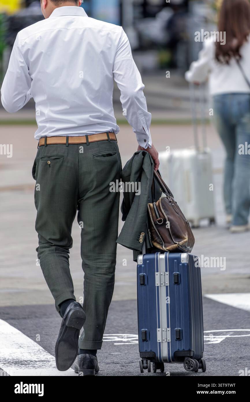 Ein Geschäftsmann streift über eine städtische plaza und überprüft das Smartphone, während er gleichzeitig mit einer Hand seine Jacke und Tasche greift. Stockfoto