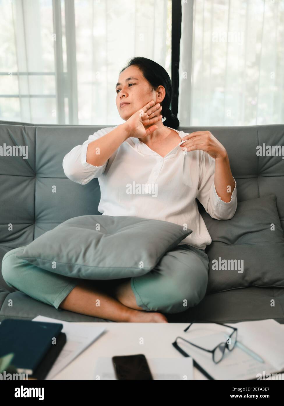 Asiatische Frau, die zu Hause mit Nackenbeschwerden auf einem Sofa sitzt, ihre Schulter in der Nähe von Papieren und Brillen berührt, zeigt Büromüdigkeit, schlechte Haltung, St Stockfoto