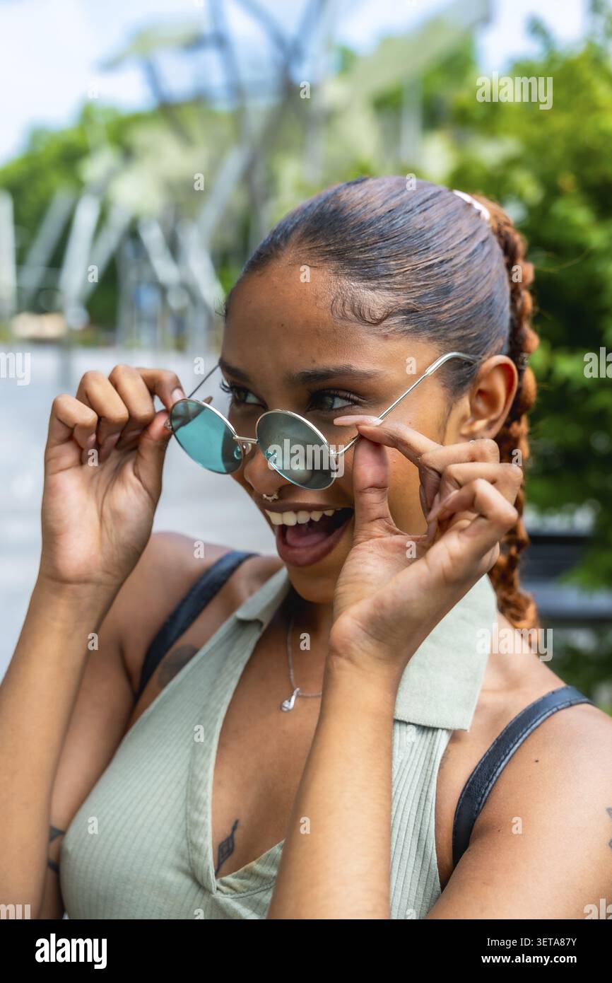 Birassische junge Frau, die eine runde Sonnenbrille senkt, verspielt mit einem breiten Lächeln blickt und Glück und Freude ausdrückt, während sie einen lebhaften Sommertag genießt Stockfoto
