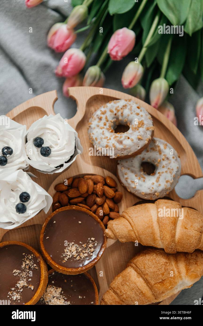 Ein wunderschöner Hochwinkel eines hölzernen Serviertabletts gefüllt mit köstlichen Leckereien, einschließlich glasierter Donuts, Schokoladentörtchen, frischen Croissants, Stockfoto