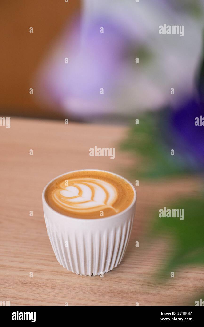 Ein frisch gebrühter heißer Cappuccino mit detaillierter Latte-Kunst, serviert in einer weißen gerippten Keramikschale auf einem natürlichen Holztisch. Stockfoto