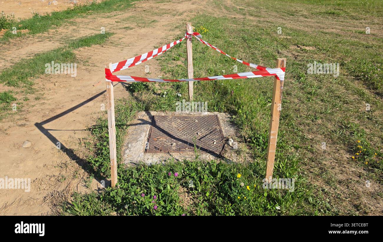 Ein Deckel aus Metall auf einem Betonuntergrund ist von einem temporären Sicherheitsband aus gestreiftem Band und Holzpfählen in einem grasbewachsenen Gelände umgeben Stockfoto