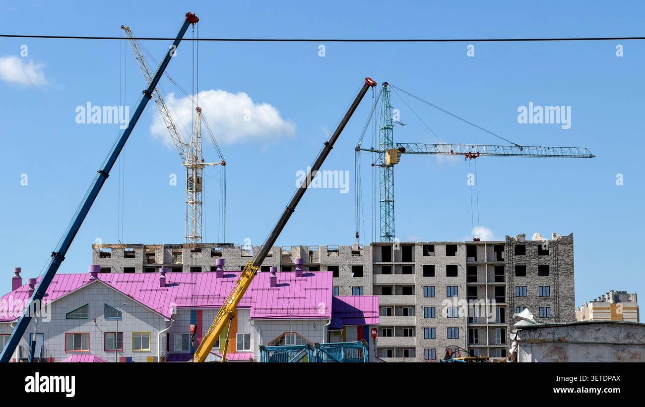 Mehrere Baukräne arbeiten auf der Baustelle eines modernen mehrstöckigen Wohnhauses unter klarem blauem Himmel. Stockfoto
