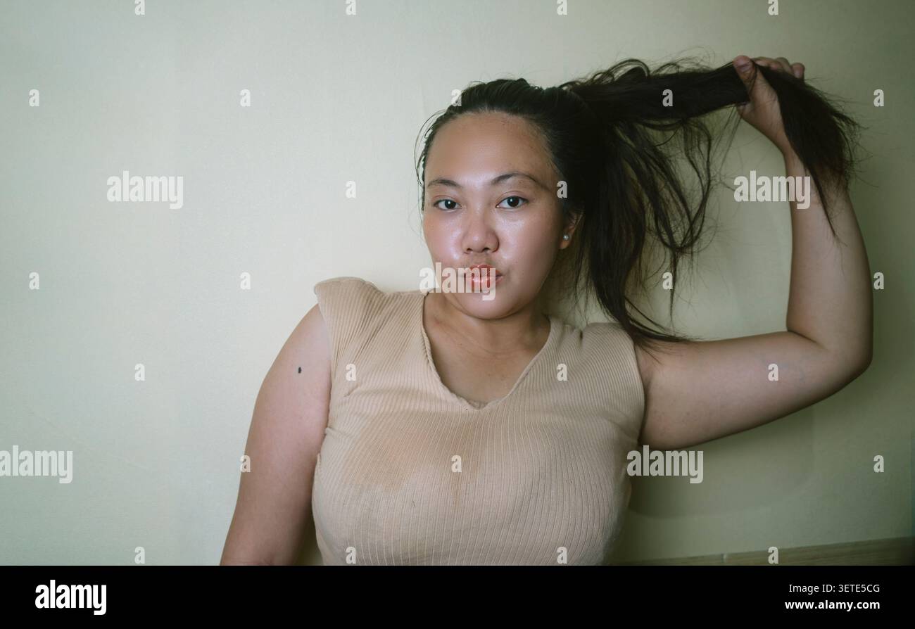 Junge Frau, die ihr langes Haar hält, während sie drinnen auf die Kamera blickt, zeigt natürliche Schönheit, persönliche Pflege, gesundes Haar. Stockfoto