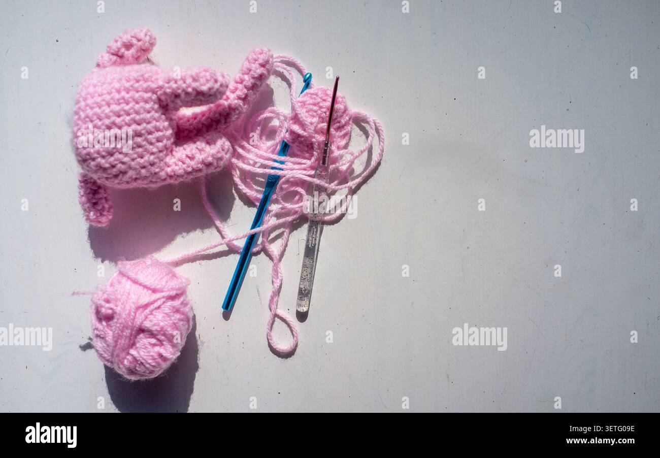 Weiche rosafarbene Babyartikel mit Garnschlaufen, Metallhaken und gestrickten Textilstücken auf einer sauberen weißen Oberfläche mit sanfter Seitenbeleuchtung Stockfoto