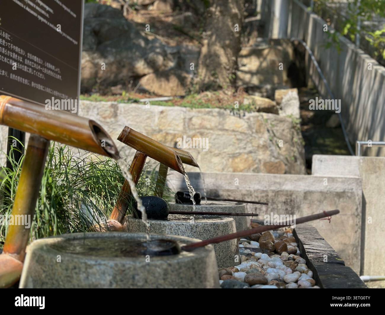 Nahaufnahme eines Bambusbrunnens mit Steinbecken zur rituellen Reinigung. Klares Wasser fließt aus traditionellen Bambusrohren in Steinschalen mit langen Stockfoto