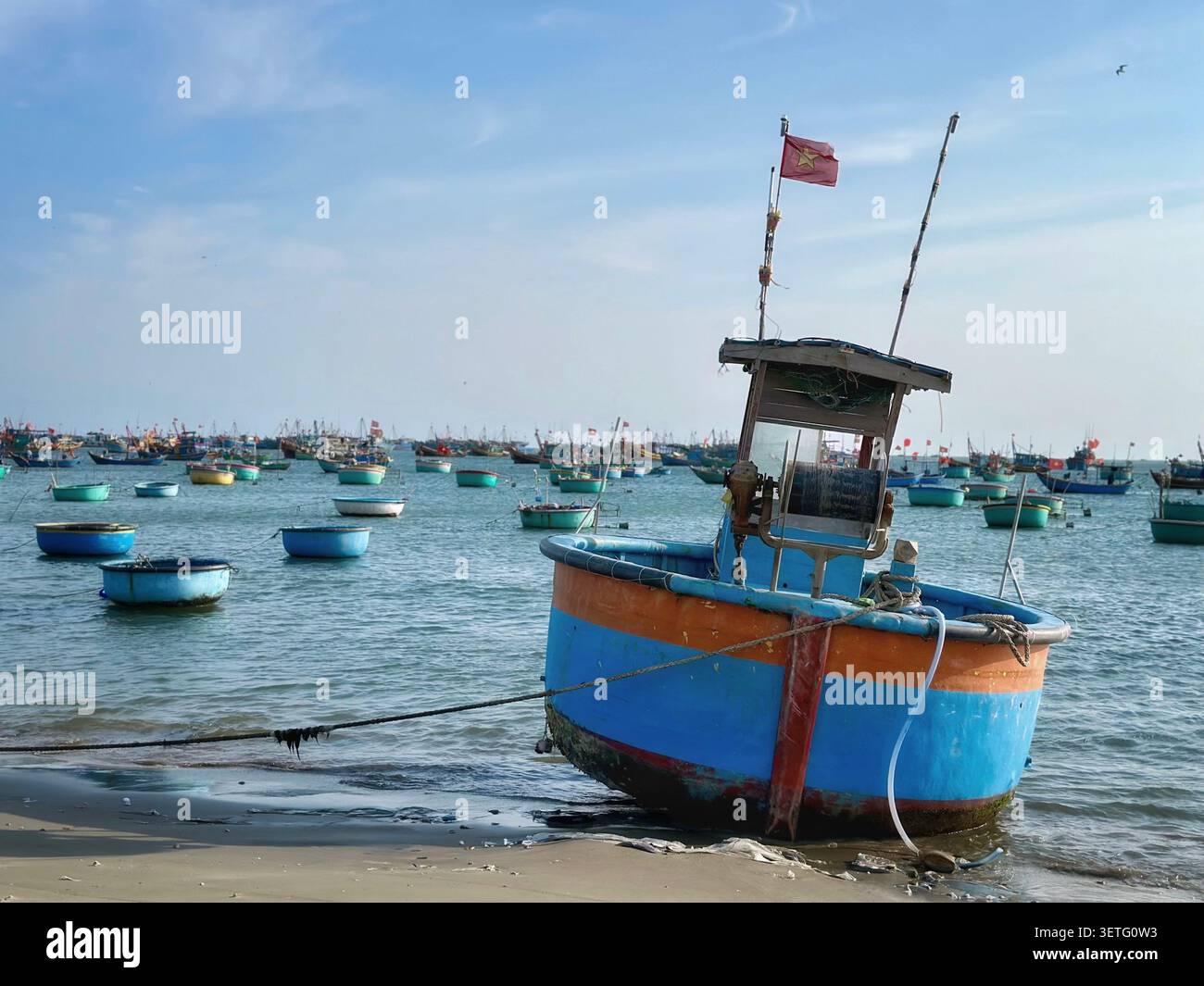 Ein leuchtend blau-orange motorisiertes Korakelboot liegt am Strand. Im Hintergrund schweben Dutzende traditioneller Fischereifahrzeuge in der ruhigen Bucht wa Stockfoto