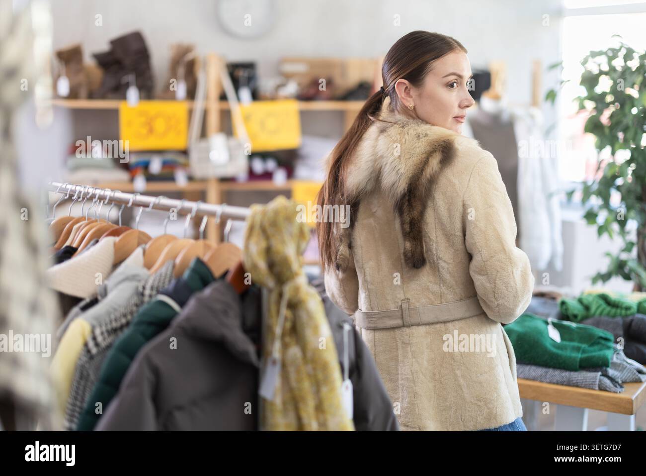 Schöne Frau, die im Bekleidungsgeschäft steht und einen neuen warmen Pelzmantel am Black Friday-Verkauf der Winterbekleidungskollektion ausprobiert Stockfoto