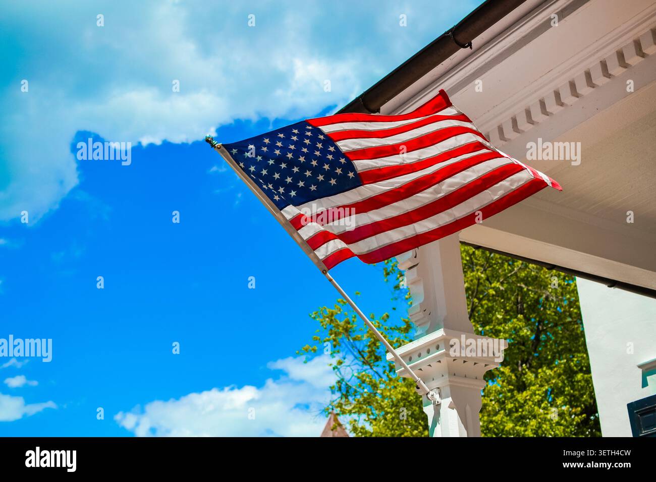 Lehigh County, Pennsylvania, USA – 19. Juni 2026: Flagge der Vereinigten Staaten, die an einem sonnigen Sommertag von der Veranda eines weißen Hauses weht. Stockfoto