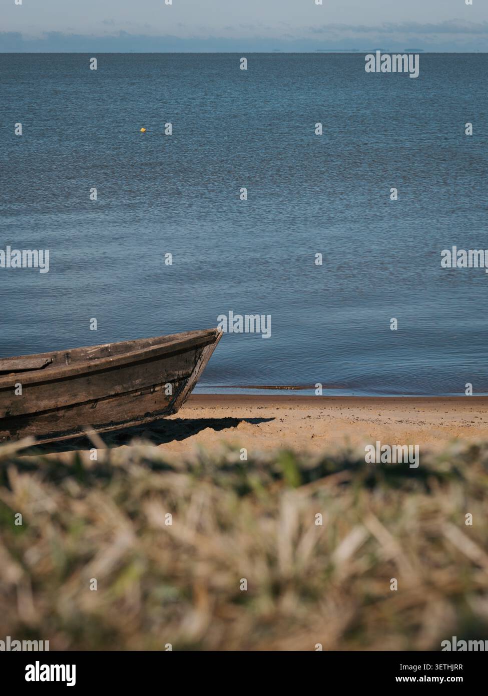 Verwittertes Holzboot ruht auf Sandstrand. Ruhiges blaues Wasser zieht sich in Richtung entfernter Hügel. Sonnenlicht leuchtet sanft auf die Wasseroberfläche. Getrocknetes Gras f Stockfoto