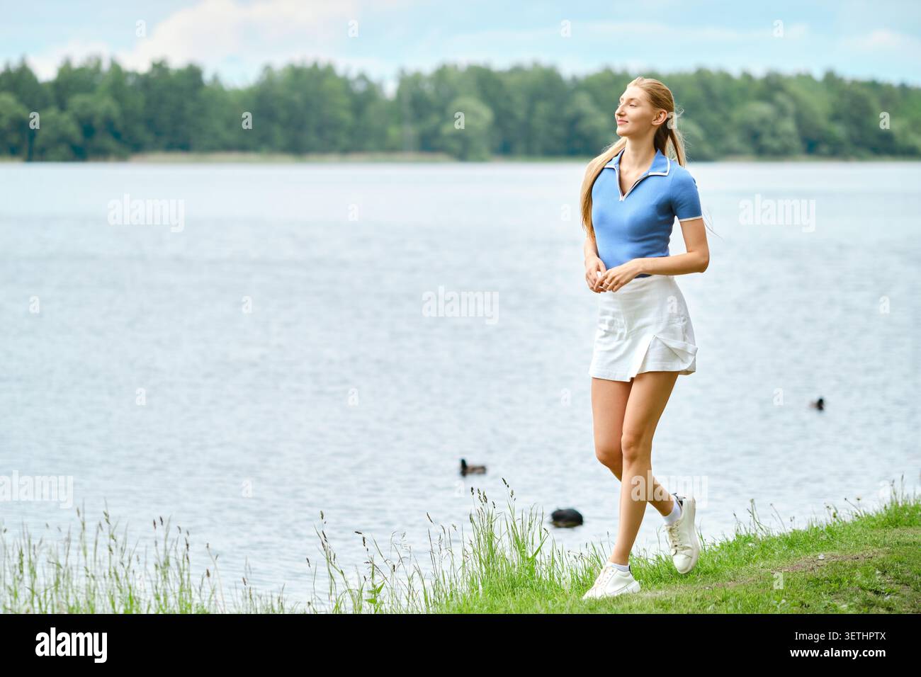 Eine Frau läuft am Ufer des Sees entlang und trägt ein blaues Hemd und weiße Shorts. Das Wasser ist ruhig, die Bäume sind grün und in der Nähe schwimmen Enten. Es ist sonniger Tag mit wenig cl Stockfoto