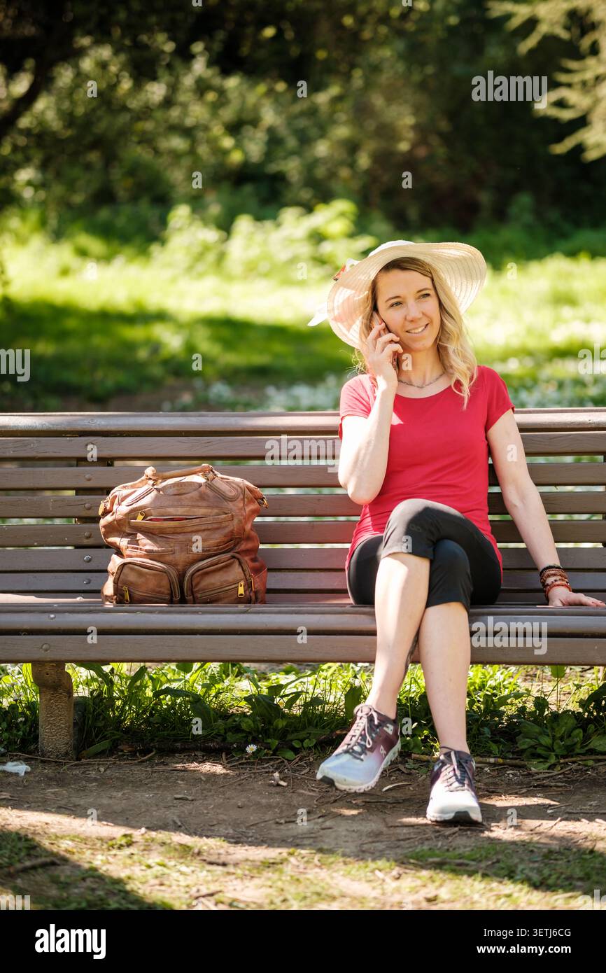Frau mit Hut, die auf der Bank im Park sitzt und mit dem Rucksack auf dem Smartphone spricht Stockfoto
