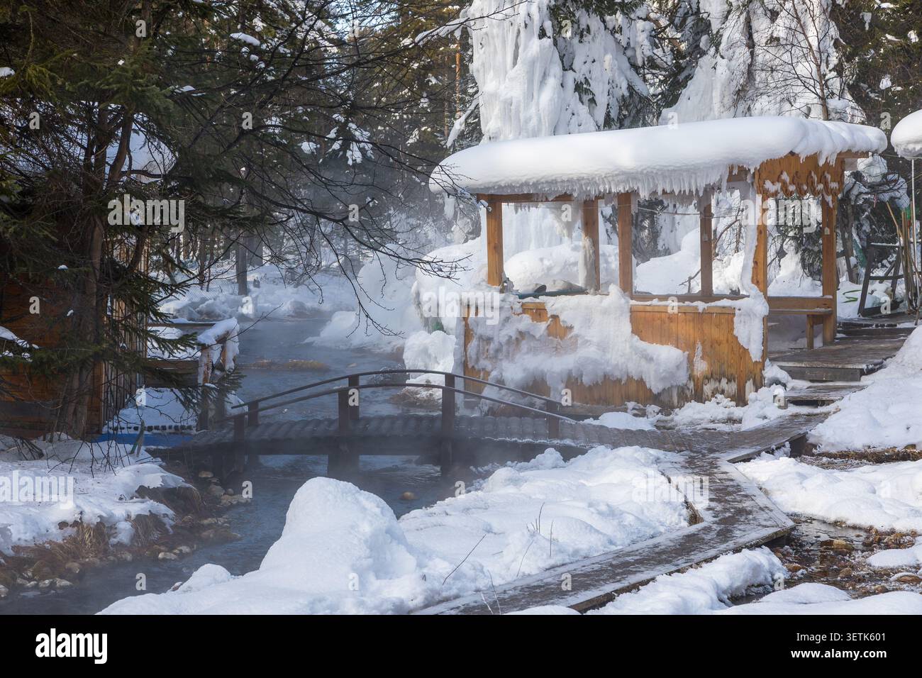 Blick auf Goryachiye Istochniki Khakusy mit einer Holzbrücke über eine dampfende Thermalquelle, schneebedeckten Bäumen und einem hölzernen Pavillon in Severobaykal'Ski Stockfoto