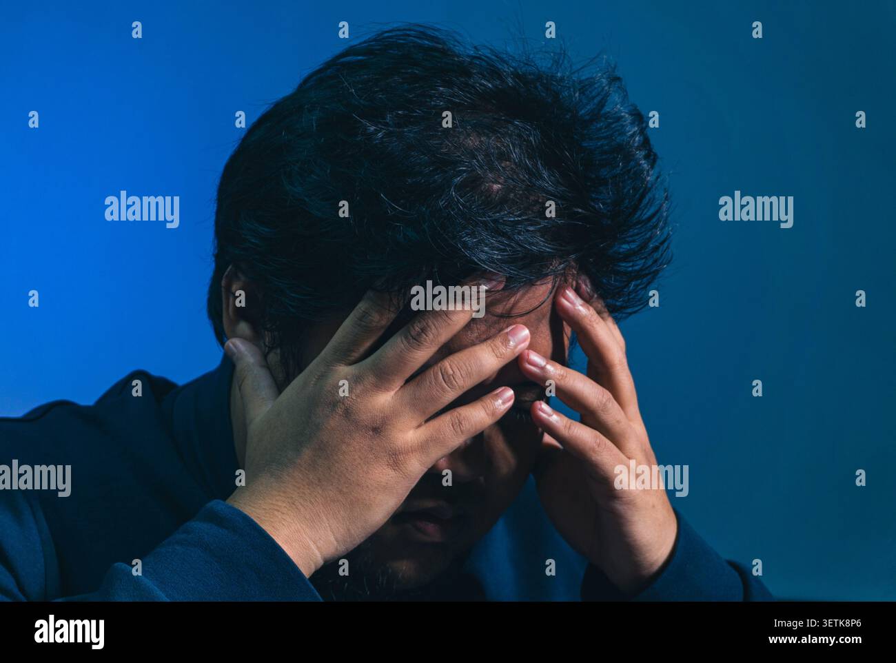 Ein überforderter erwachsener Mann bedeckt sein Gesicht mit beiden Händen in einer Geste von intensivem Stress, starker Angst und emotionaler Verzweiflung. Er schaut wie ein Heav herunter Stockfoto