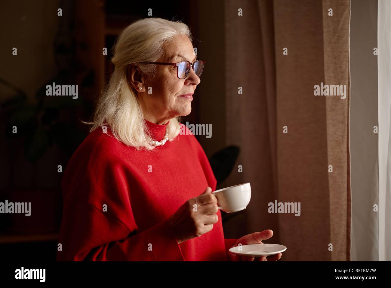 Seniorin hält Kaffeetasse und schaut zu Hause durch das Fenster Stockfoto