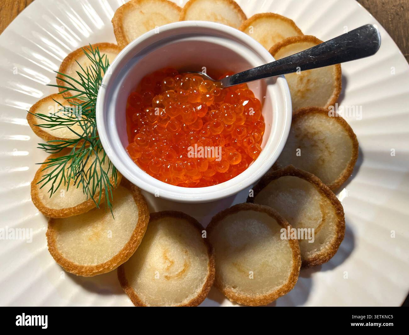 Leuchtend orange Lachsrogen Kaviar serviert in einer weißen Schüssel mit einem Löffel, umgeben von goldenen Blinis im Kreis und frischem Dill auf einem weißen Teller Stockfoto