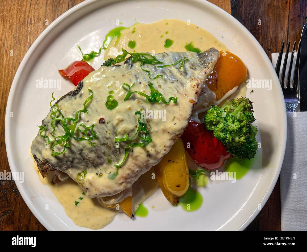 Weißer Teller mit gegrilltem Branzino Seebarsch-Fischfilet mit cremiger Kräutersauce, begleitet von geröstetem Gemüse. Stockfoto
