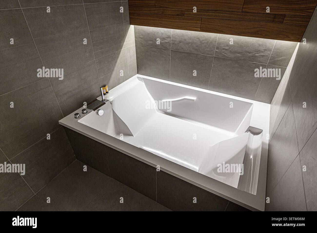 Das moderne Bad verfügt über eine elegante, weiße Badewanne mit dunkelgrauen Fliesen. Holzakzente und dezente Beleuchtung schaffen ein Spa-ähnliches Ambiente Stockfoto
