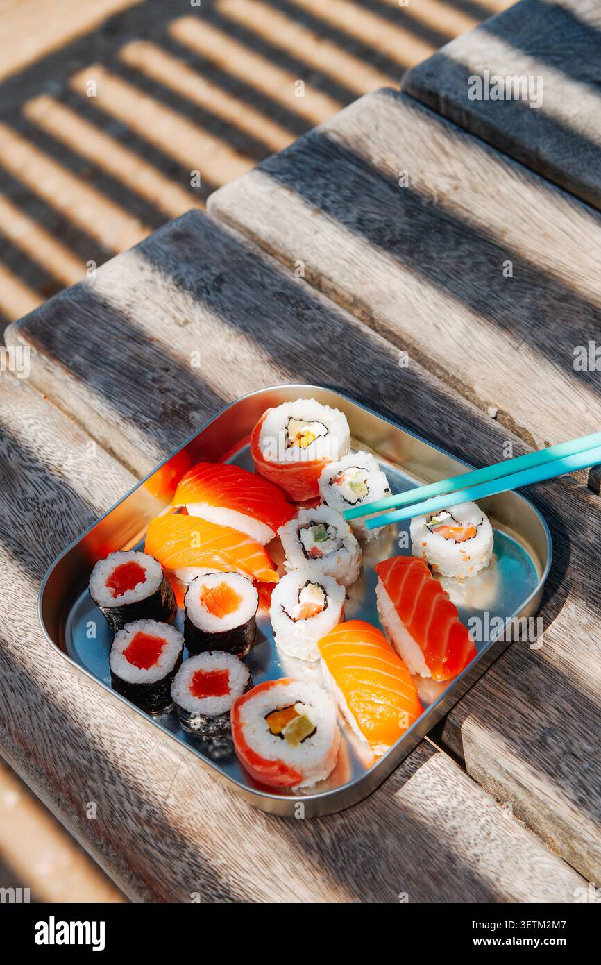 Ein mit Sushi gefüllter Edelstahlbehälter steht auf einer Holzbank im Freien, mit türkisfarbenen Essstäbchen Stockfoto