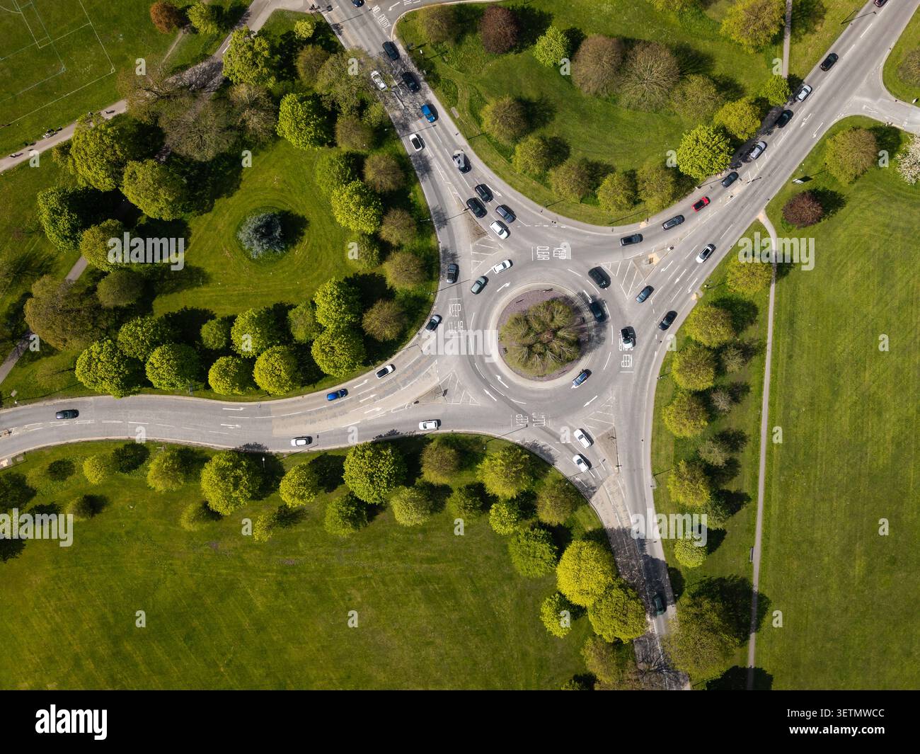Aus der Vogelperspektive direkt über einem von Bäumen gesäumten Verkehrskreis oder Kreisverkehr mit Autos und Fahrzeugen in Bewegung zur Hauptverkehrszeit durch die Landschaft mit grünem Gras Stockfoto