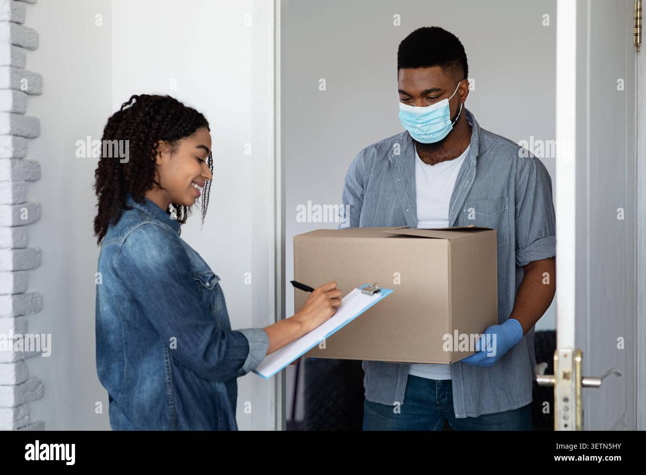 Sichere Lieferung Nach Hause. Schwarze Frau Erhielt Parcel Von Courier In Medizinischer Maske Stockfoto