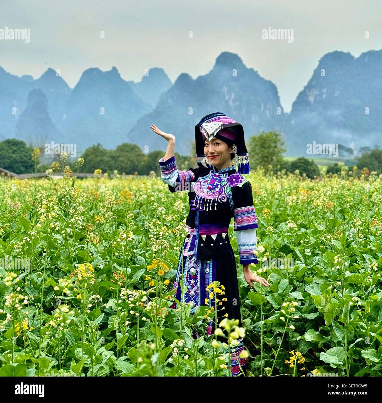 Frau in traditionellem, besticktem Kostüm der ethnischen Minderheit, tanzt im Rapsblumenfeld mit Karstbergen, Guangxi, China - Smartphone-aufgenommenes Stockfoto