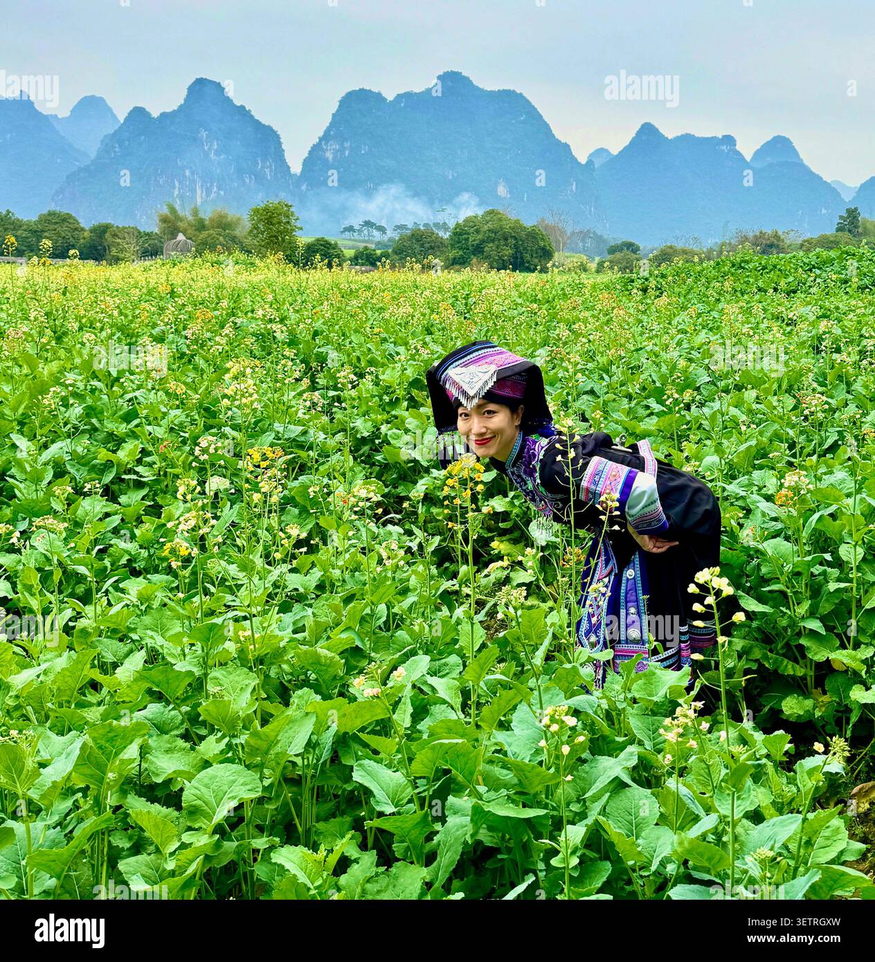 Lächelnde Frau im traditionellen Kopfschmuck der ethnischen Minderheit, die durch Rapsblumenfeld mit Karstbergen in Guangxi, China blickt - Smartphone-aufgenommenes Stockfoto