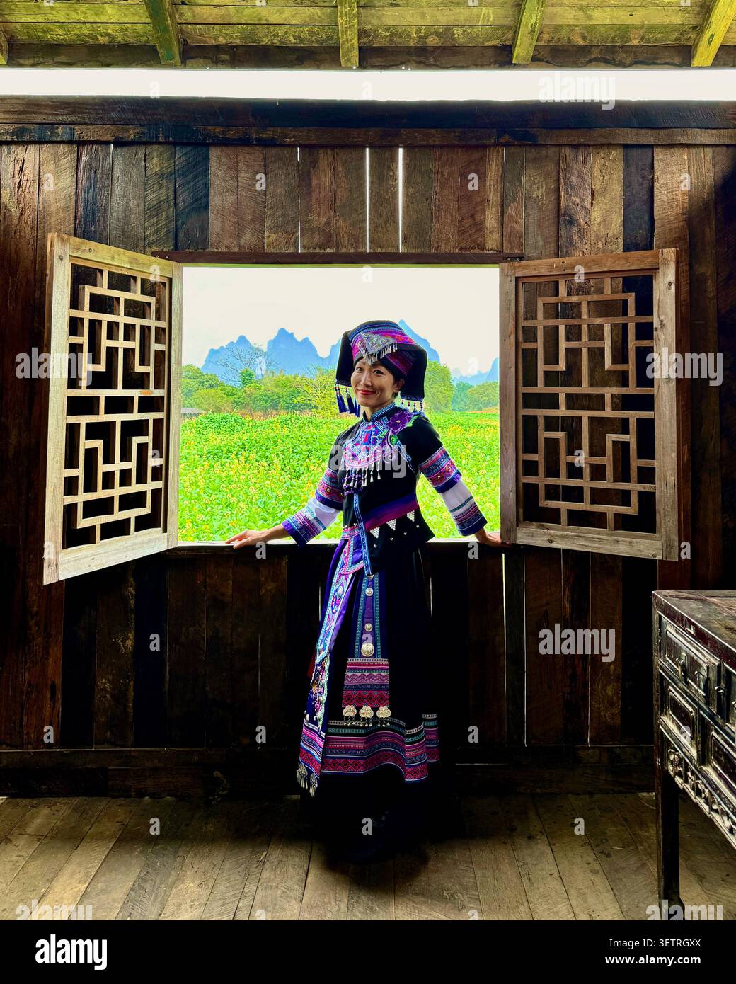 Frau in traditioneller Tracht ethnischer Minderheiten, die an einem offenen traditionellen Gitterfenster mit grünem Feld und Karstbergen steht, ländliches Guangxi, China - Smartphone-aufgenommenes Stockfoto