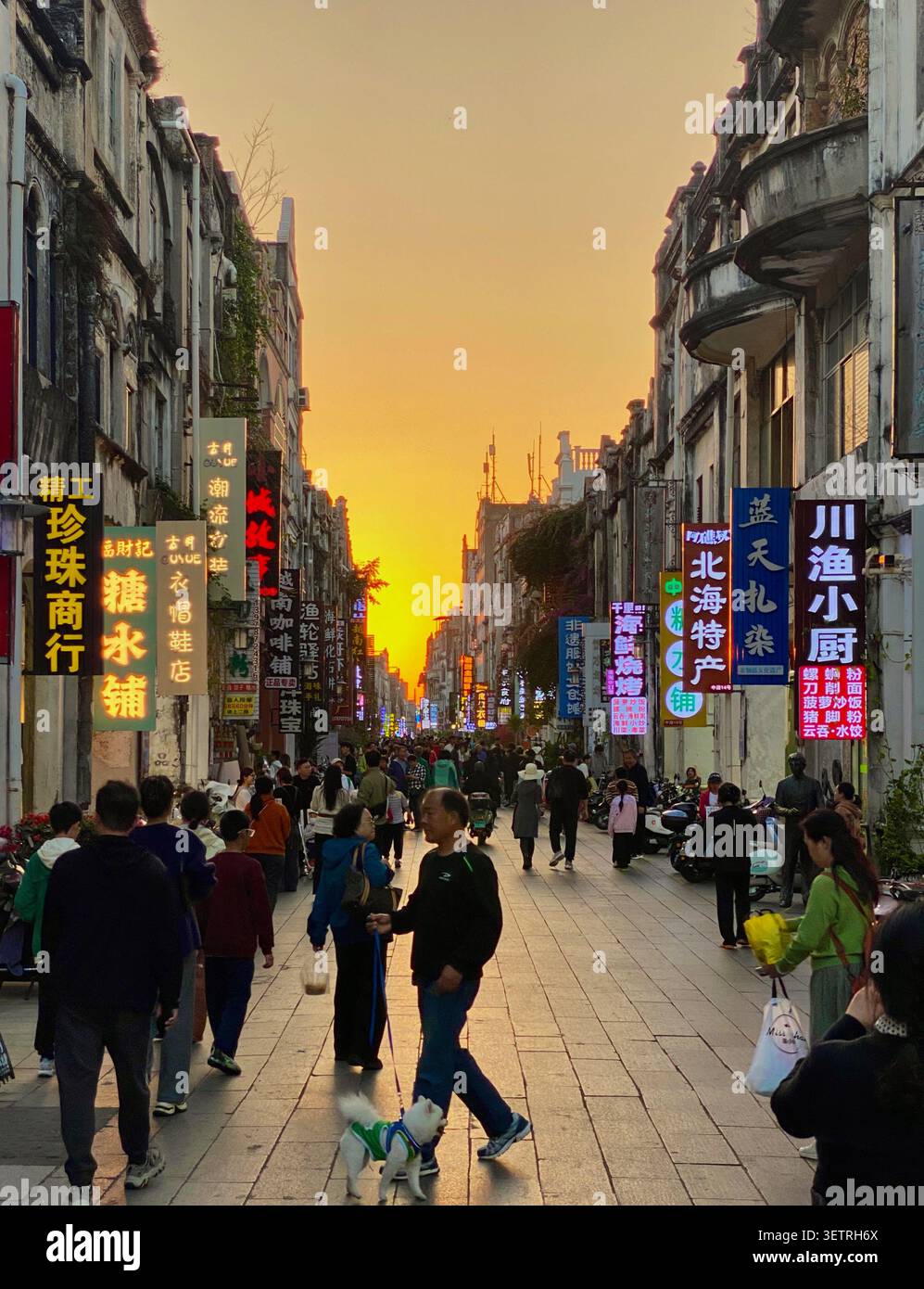 Chinesische Fußgänger-Einkaufsstraße bei dramatischem Sonnenuntergang mit leuchtenden Neonreklamen und orangefarbenem Himmel, Beihai Old Street, Guangxi, China - Smartphone-aufgenommenes Stockfoto