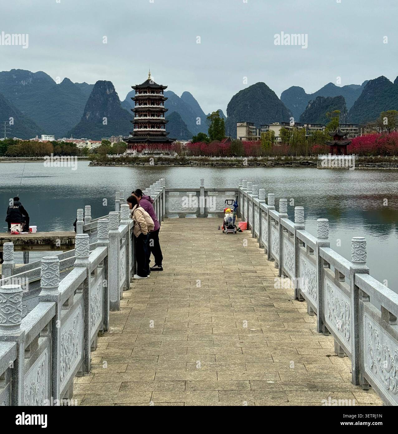 Asiatisches urbanes Leben eines Paares über einer traditionellen Brücke, beobachten einen Mann beim Angeln, mit einer historischen Pagode und Karstbergen im Hintergrund - Smartphone-aufgenommenes Stockfoto