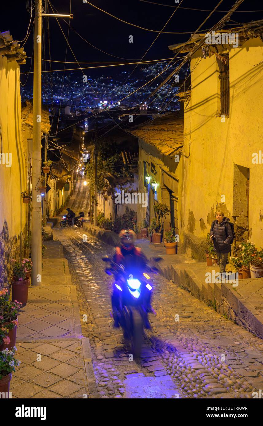 Biker und ein Fußgänger in einer steilen, engen gepflasterten Straße in Cusco, Peru, in der späten Nacht, mit den Lichtern der Stadt im Hintergrund. Stockfoto