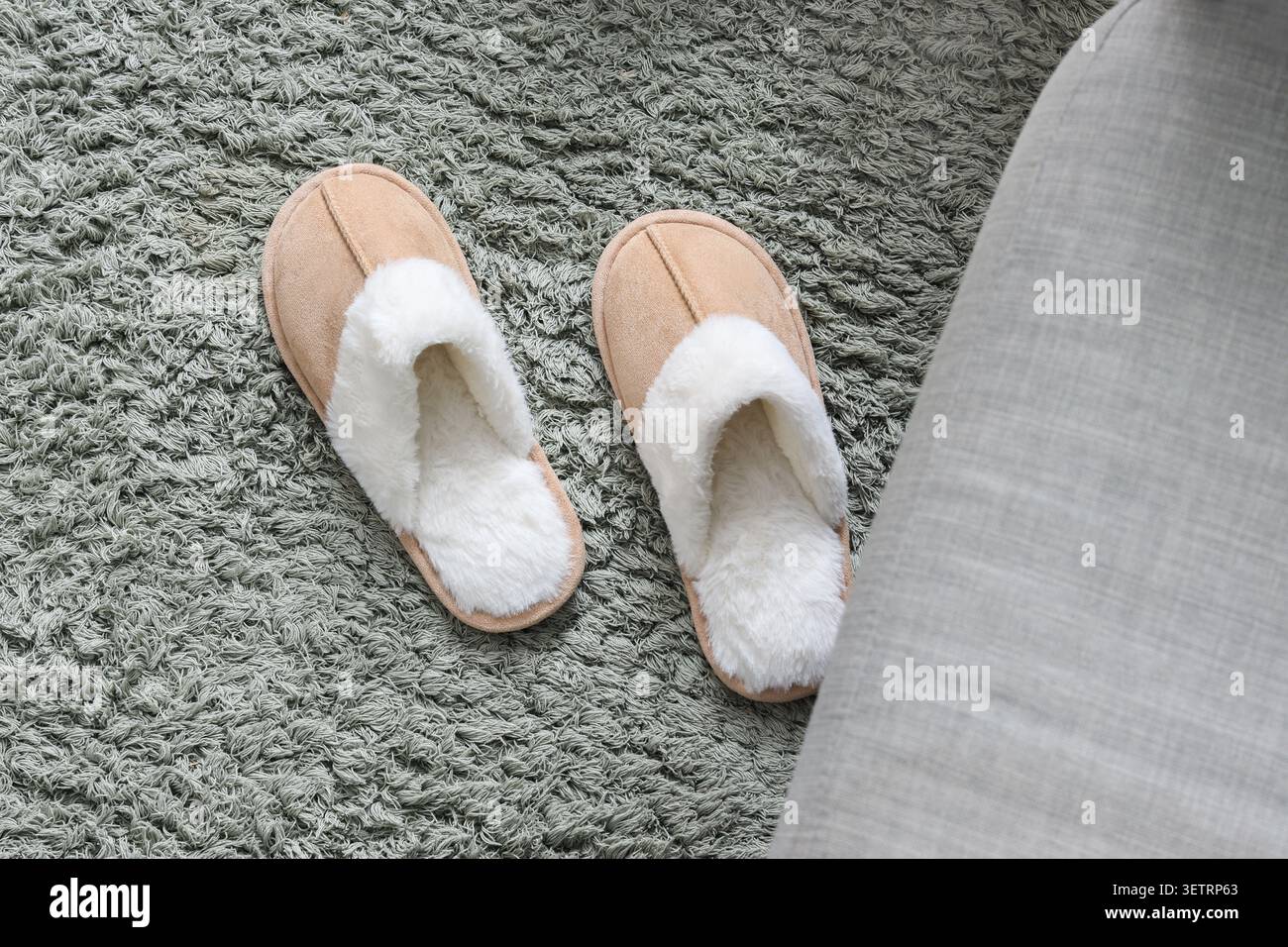 Zwei gemütliche beige Hausschuhe auf einem strukturierten grauen Teppich neben einem grauen Sofa Stockfoto