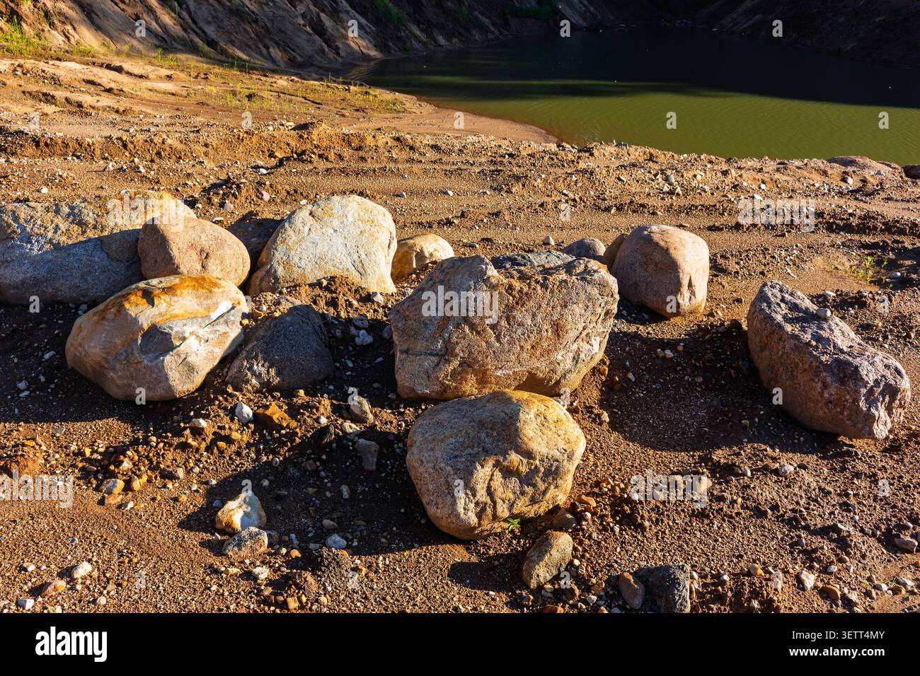Große Felsbrocken auf sandigem Boden in einem Steinbruch mit einem kleinen See im Hintergrund unter hellem natürlichem Sonnenlicht, die strukturierten Schatten erzeugen Stockfoto